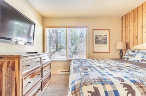 Une chambre à coucher comprenant un lit, une commode, un téléviseur, une fenêtre avec des stores et un tableau encadré accroché au mur.