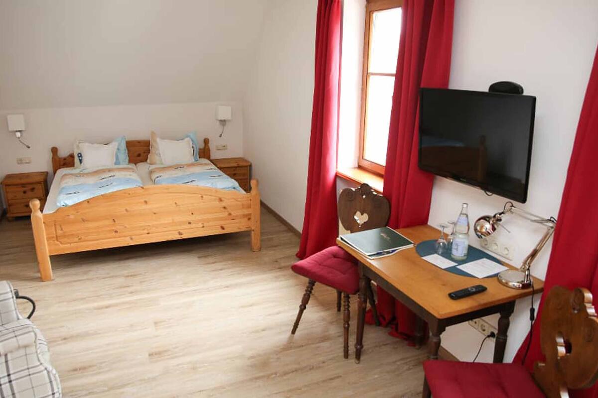 A bedroom with a wooden bed, a desk with a chair, a television, and a window with red curtains.