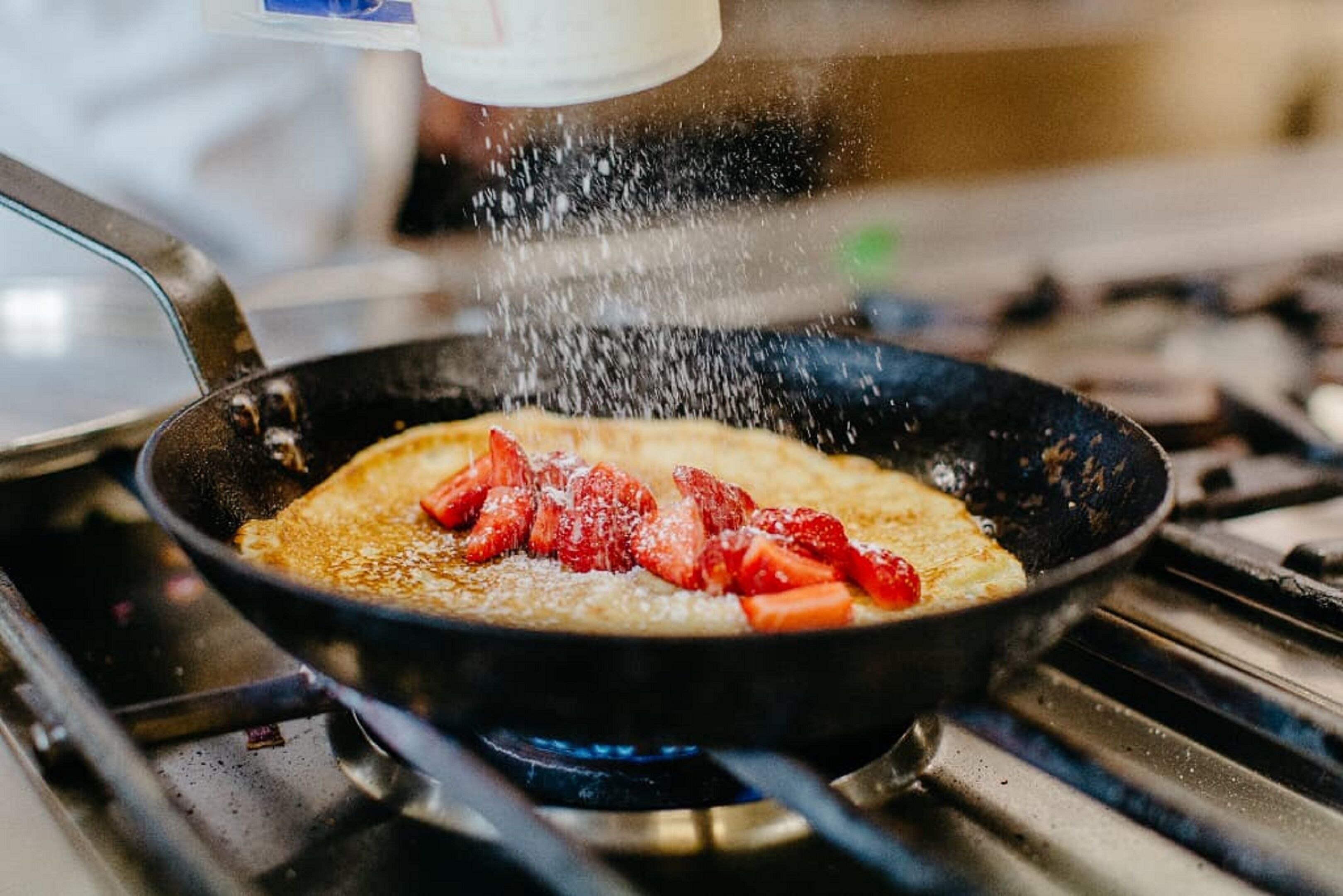 Une crêpe en train de cuire dans une poêle, garnie de fraises et saupoudrée de sucre glace.