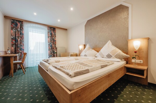 Une chambre d’hôtel avec un lit en bois, un bureau, une chaise, une table de chevet avec une lampe et une fenêtre avec des rideaux.