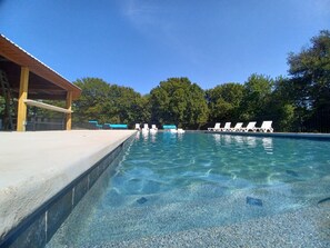 A clear swimming pool with white lounge chairs and a covered area with a wooden roof. - Tiny House Cabins (Whitewright)