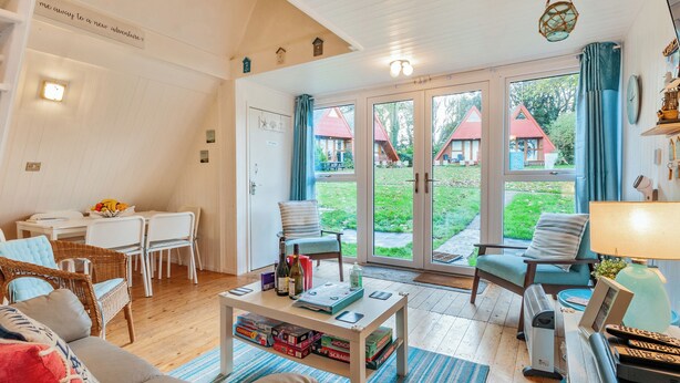 Una sala de estar acogedora con un sofá, sillas, una mesa baja con libros y botellas, un televisor y un ventilador. Hay un comedor con una mesa y sillas, y una vista de un jardín con un edificio de techo rojo.