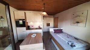 A compact room with a kitchenette, a wooden dining table, a bed with a floral bedspread, and a ceiling light. - Haus Naturenergie (Frauenau)