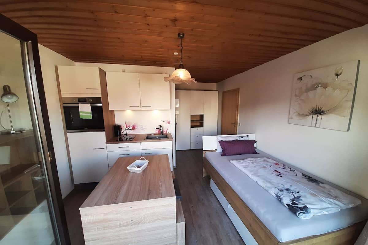 A compact room with a kitchenette, a wooden dining table, a bed with a floral bedspread, and a ceiling light.