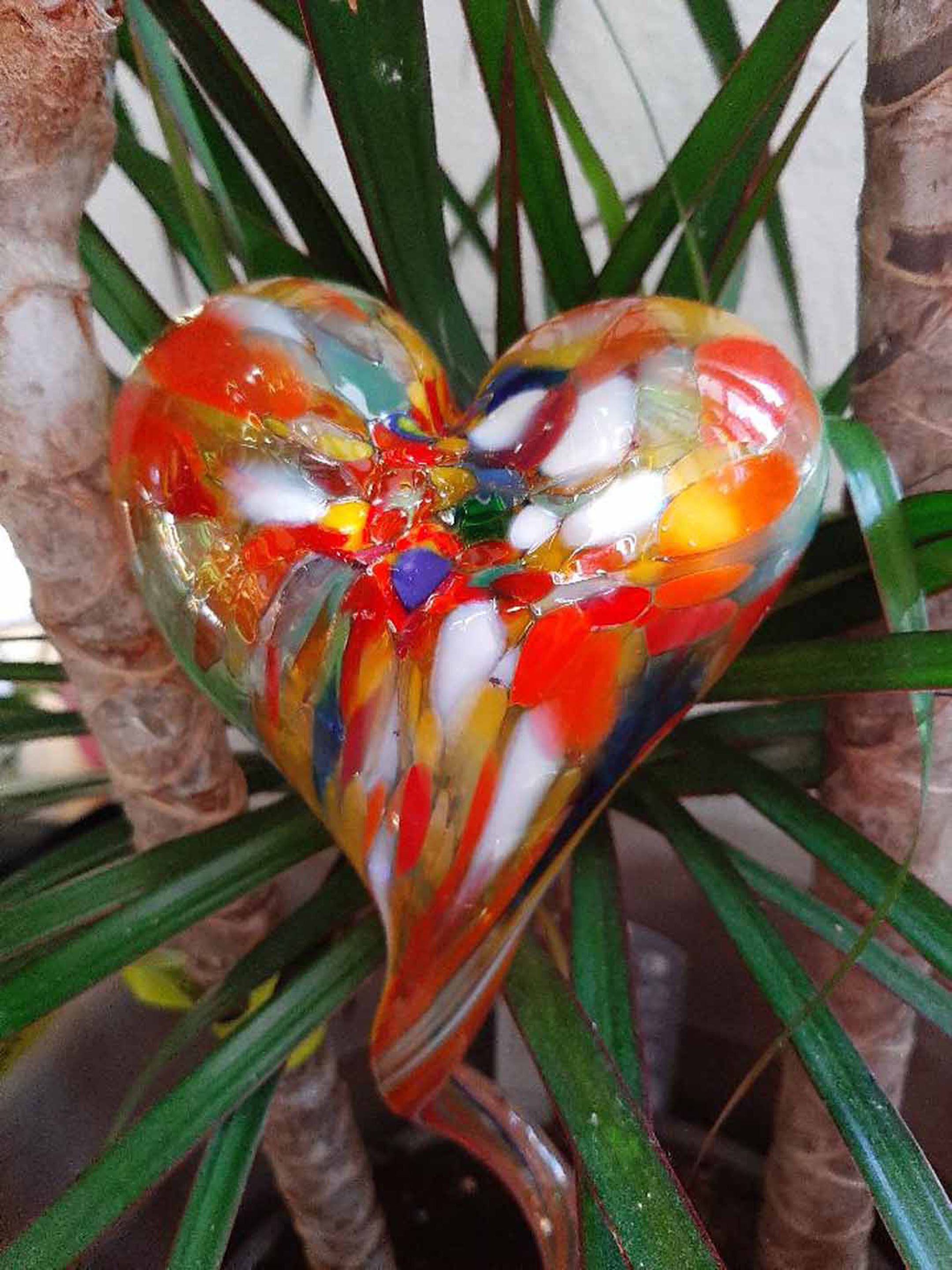 A heart-shaped glass ornament with multicolored glass pieces, hanging amidst green leaves.