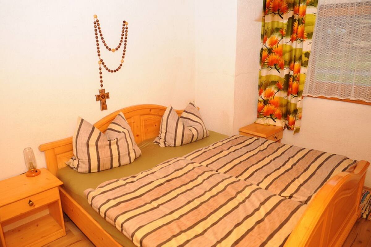 A single-bed room with a wooden headboard, striped bedding, a nightstand with a lamp, and a wall-mounted cross and rosary.