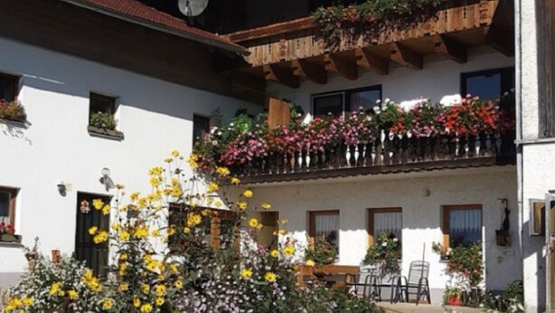 A courtyard with a white building, a wooden balcony with flowers, and a table with chairs. - Woferlhof Ferienhof Boxhorn (Böbrach)