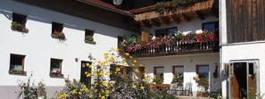 Une cour avec un bâtiment blanc, un balcon en bois orné de fleurs et une table entourée de chaises.