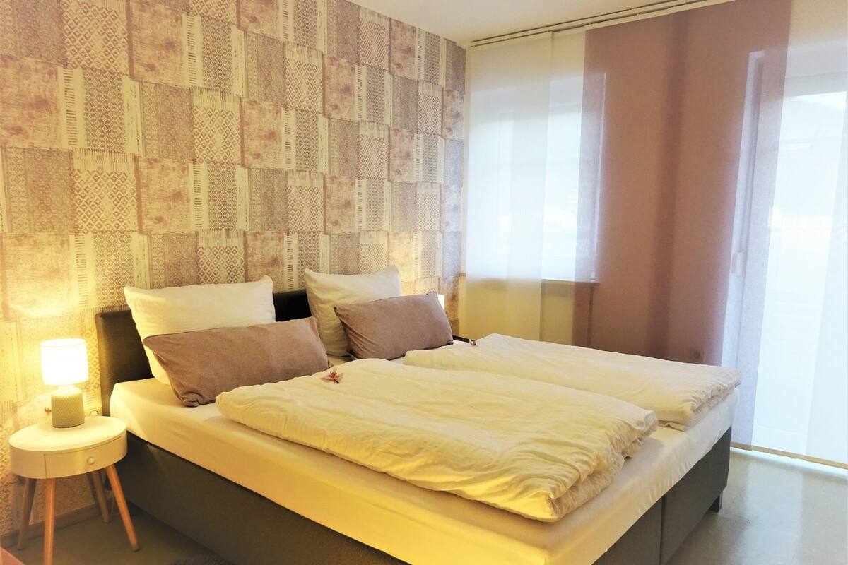 A modern hotel room with a large bed, a bedside table with a lamp, and a patterned wallpaper on the wall.
