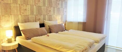 A modern hotel room with a large bed, a bedside table with a lamp, and a patterned wallpaper on the wall.