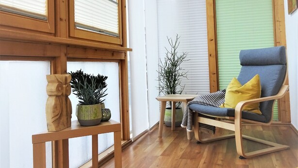Una habitación con piso de madera, ventana con marco de madera, una mesa de madera con una planta en maceta, una silla azul con cojín amarillo y un sofá gris.