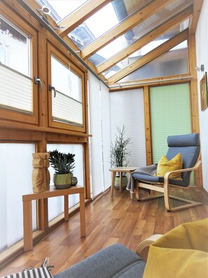 A room with wooden flooring, a wooden-framed window, a wooden table with a potted plant, a blue chair with a yellow cushion, and a grey sofa. - Ferienwohnungen Koller (Bodenmais)