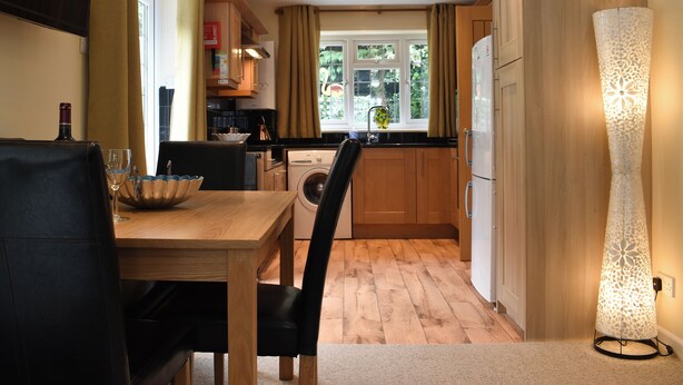 A compact kitchen with wooden cabinets, a dining table with chairs, a washing machine, and a decorative floor lamp. - 2 bedroom accommodation in Topsham, near Exeter (Topsham, near Exeter)