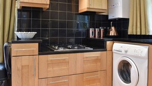 A compact kitchen with wooden cabinets, a black tiled backsplash, a stainless steel range hood, a built-in dishwasher, and a kettle on the countertop. - 2 bedroom accommodation in Topsham, near Exeter (Topsham, near Exeter)