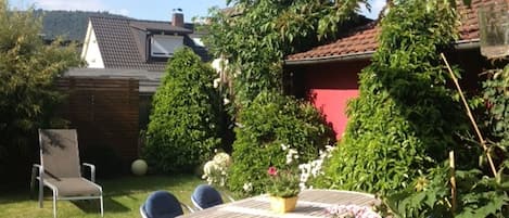 Une terrasse avec une table et des chaises, entourée de verdure, avec une maison en arrière-plan.