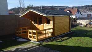 A wooden cabin with a porch and a small deck, surrounded by a grassy yard and a gravel path. - Ferienwohnung Stoiber (Frauenau)