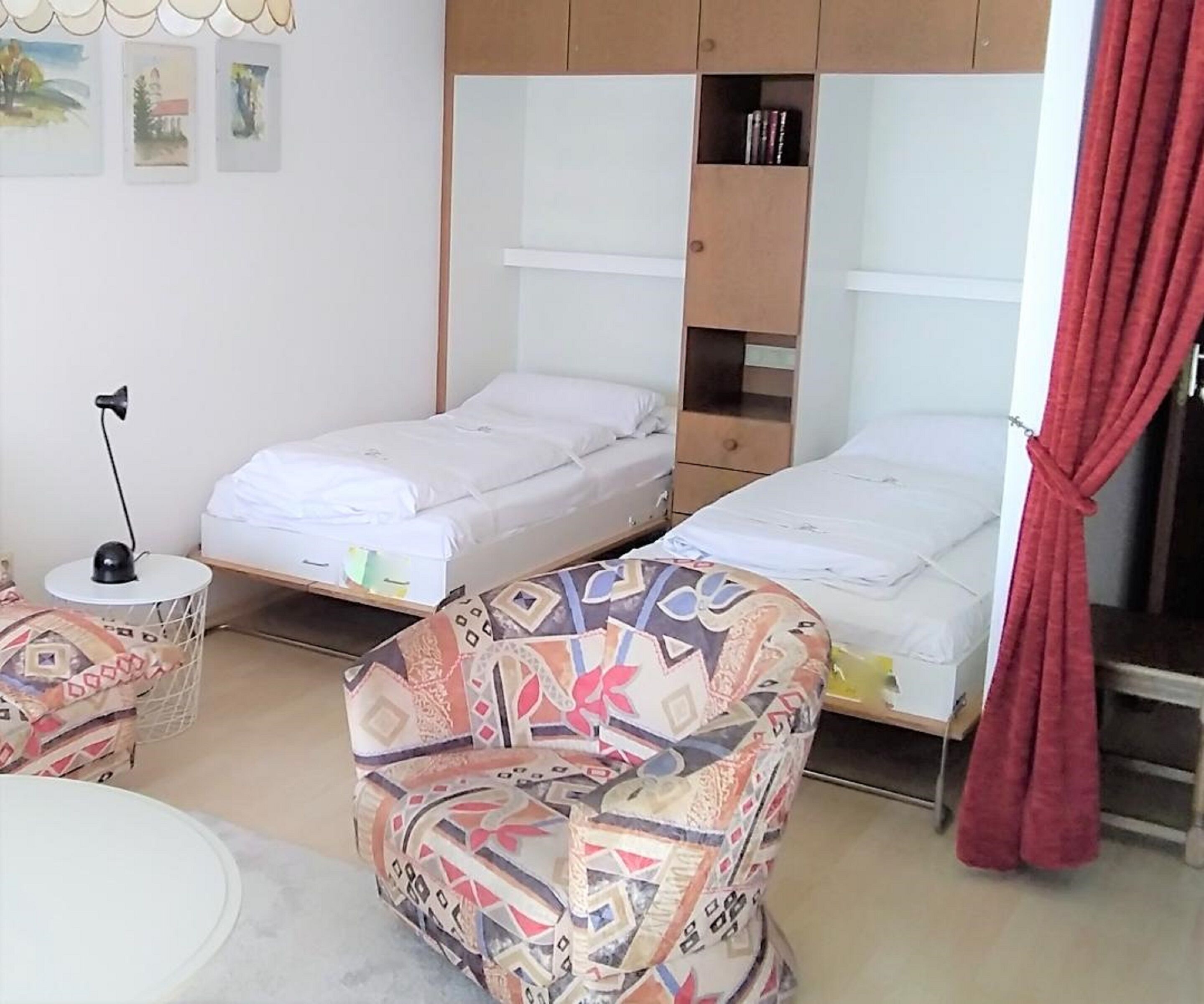 A small, compact room with two single beds, a patterned armchair, a small round table, and a desk with a lamp.