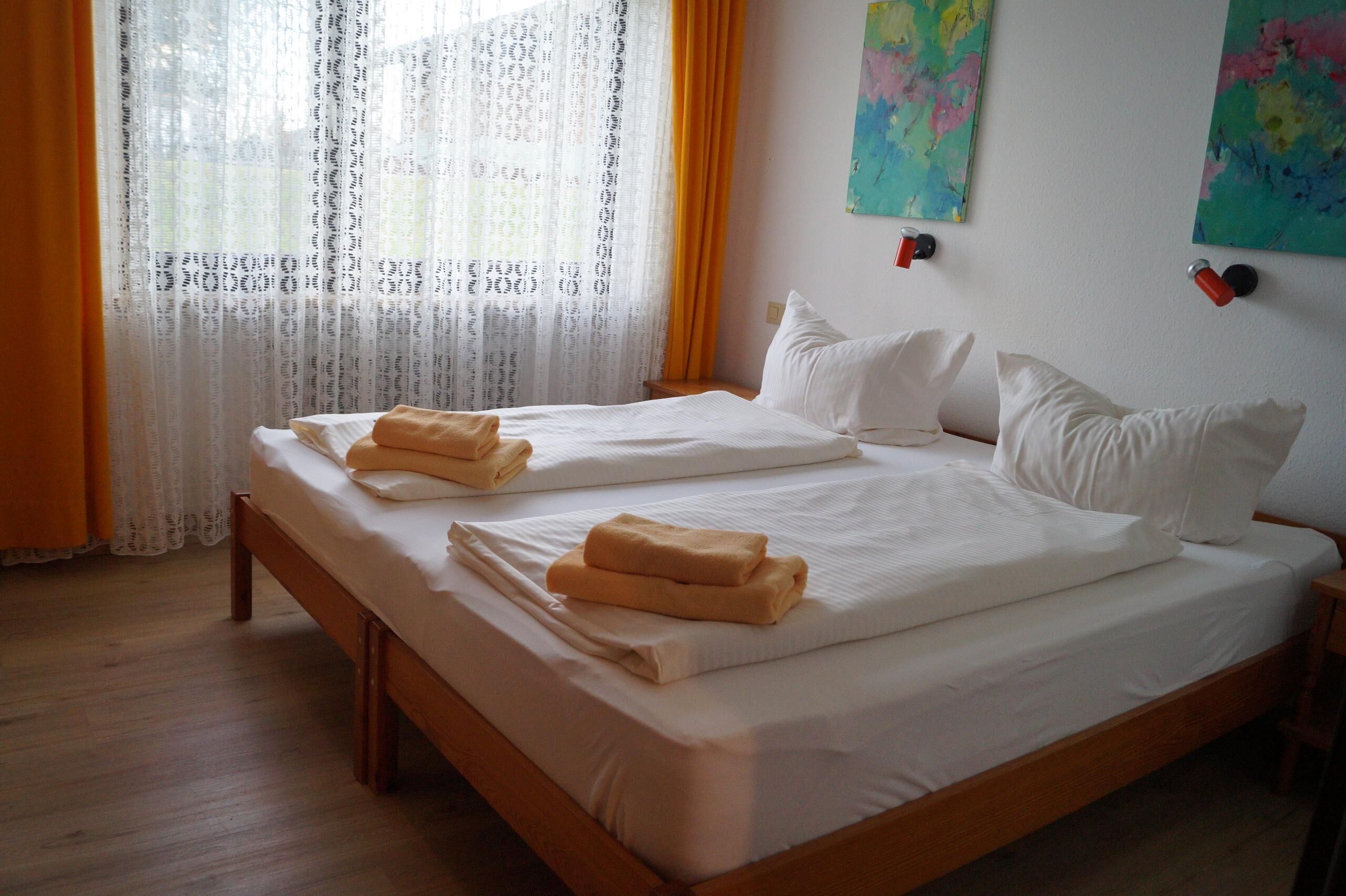 A double bed with white linens and two folded towels, a wooden headboard, and a window with sheer curtains.