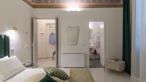 Habitación de hotel con cama, mesita de noche y un pequeño banco. Hay un espejo en la pared y un radiador debajo. La habitación tiene una puerta que da a un baño con lavabo y espejo.