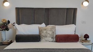 A neatly made bed with a grey headboard, white pillows, and a grey bedspread. There is a small bedside table with a vase of flowers and a book.