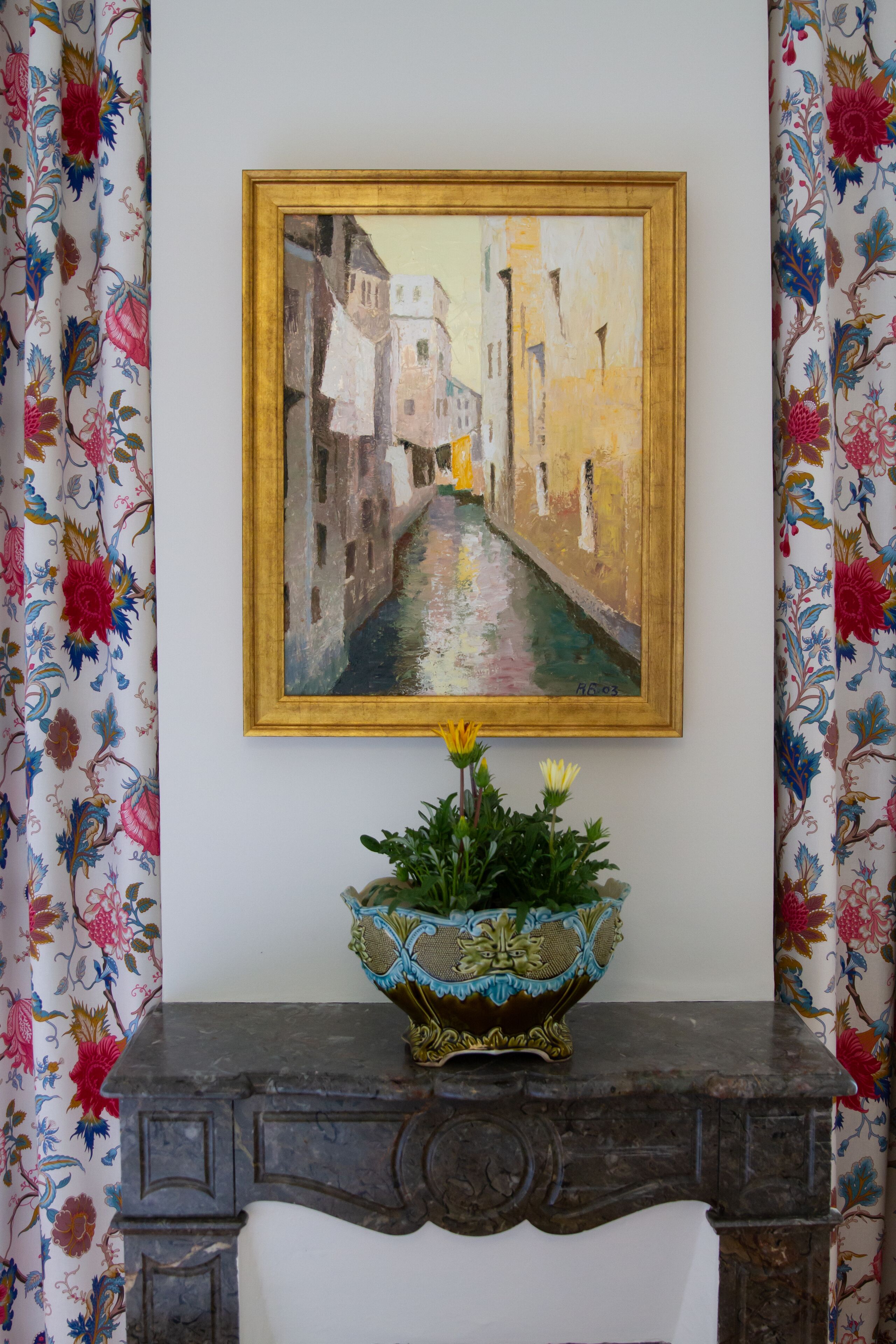 A framed painting of a canal scene on a wall, a decorative bowl on a dark wooden console table, and floral-patterned curtains.