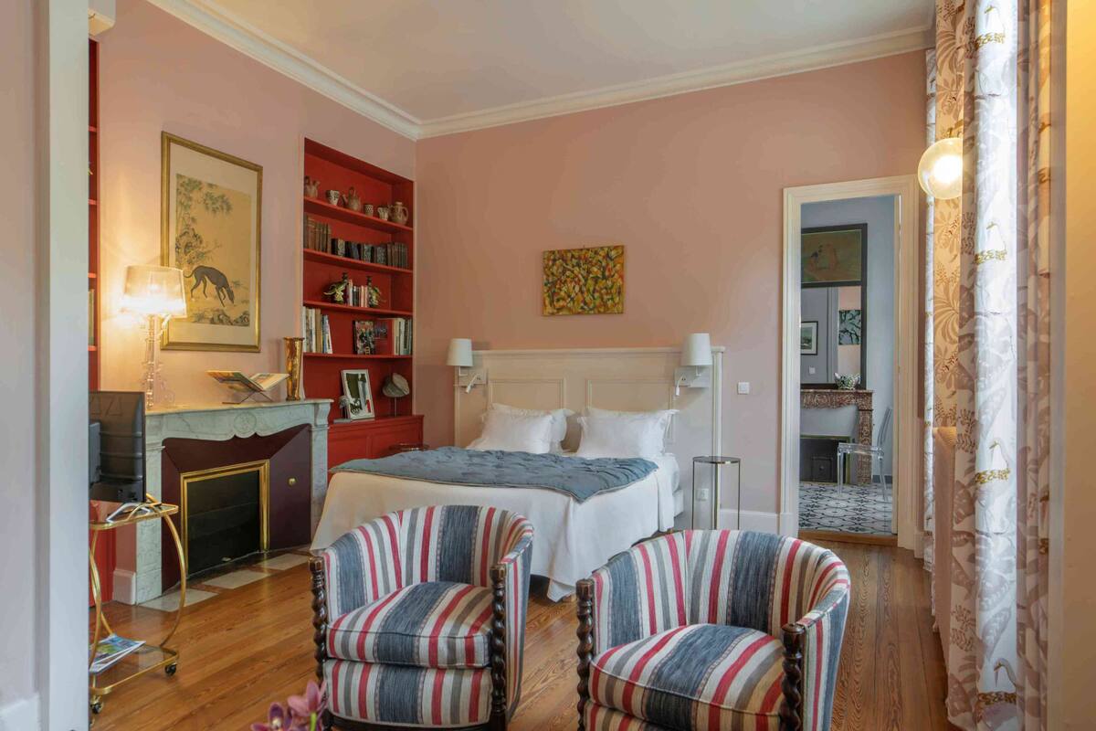 Une chambre à coucher avec un lit, deux fauteuils à rayures, une bibliothèque, une cheminée et un miroir.