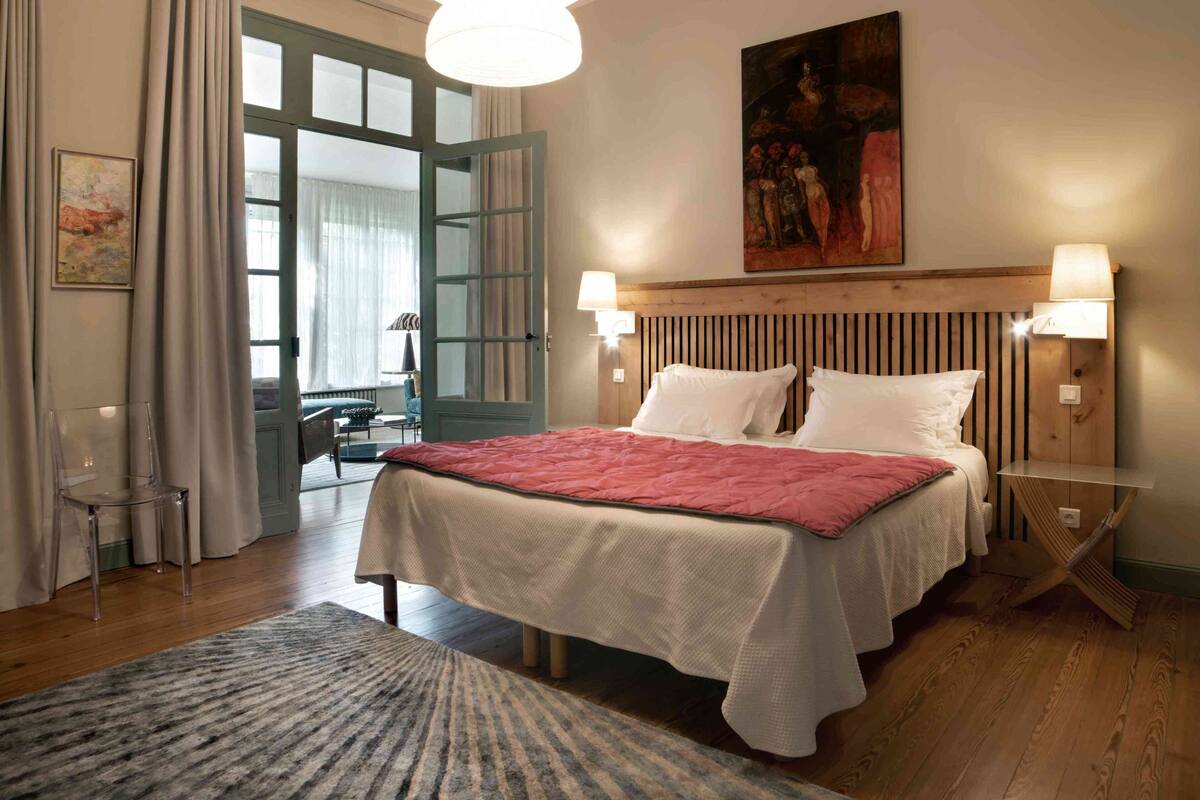 Une chambre avec un grand lit, une tête de lit en bois, deux lampes de chevet, un tableau au mur et une vue sur l’extérieur, visible à travers une porte ouverte.