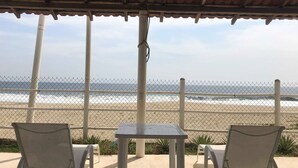 Eine Terrasse direkt am Strand mit zwei Metallstühlen und einem Tisch, mit Blick auf das Meer durch einen Zaun.