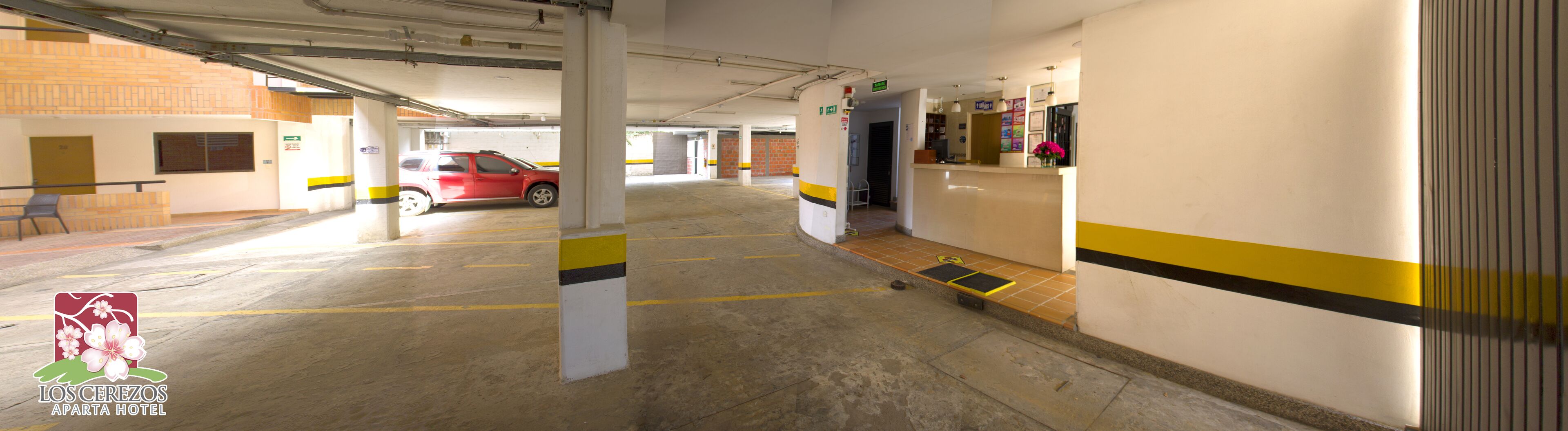 A parking garage with a red car parked in a spot, concrete pillars with yellow stripes, and a reception area with a counter and a flower vase.