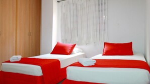 Zwei Einzelbetten mit roter Bettwäsche und weißen Kissen, ein Holzschrank und ein Fenster mit weißen Vorhängen.