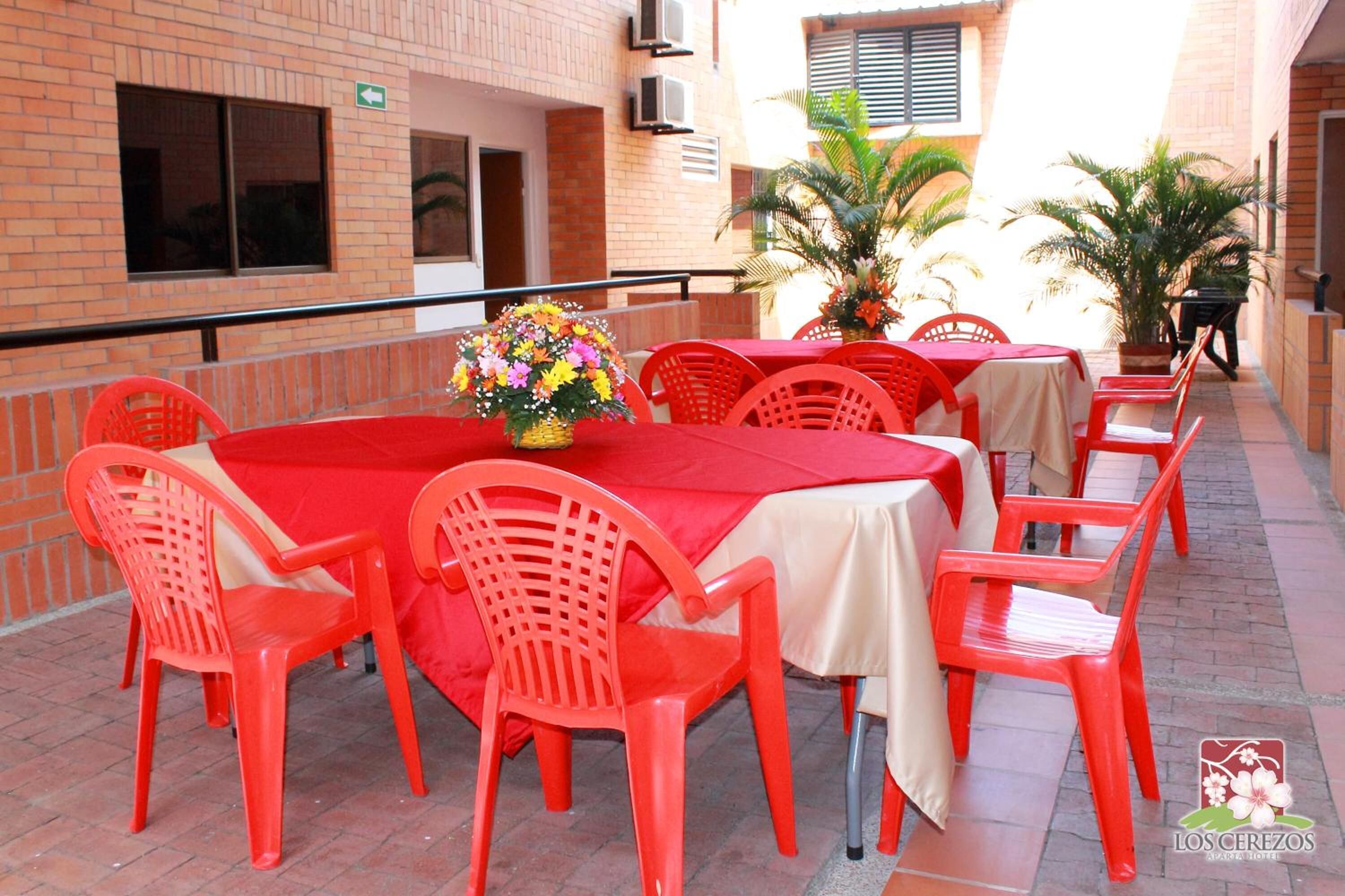 Een buitenterras met rode plastic stoelen en tafels, een bloemstuk als decoratie, en een bakstenen gebouw op de achtergrond.