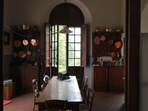 Un comedor con una mesa y sillas de madera, un ventanal con contraventanas abiertas y una colección de ollas y sartenes de cobre en estantes.