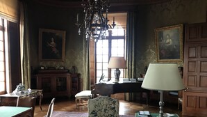 A room with a piano, a green table, a patterned armchair, a chandelier, and a large window with curtains. - Château de Joyeux - Chambres D'hôtes (Joyeux)