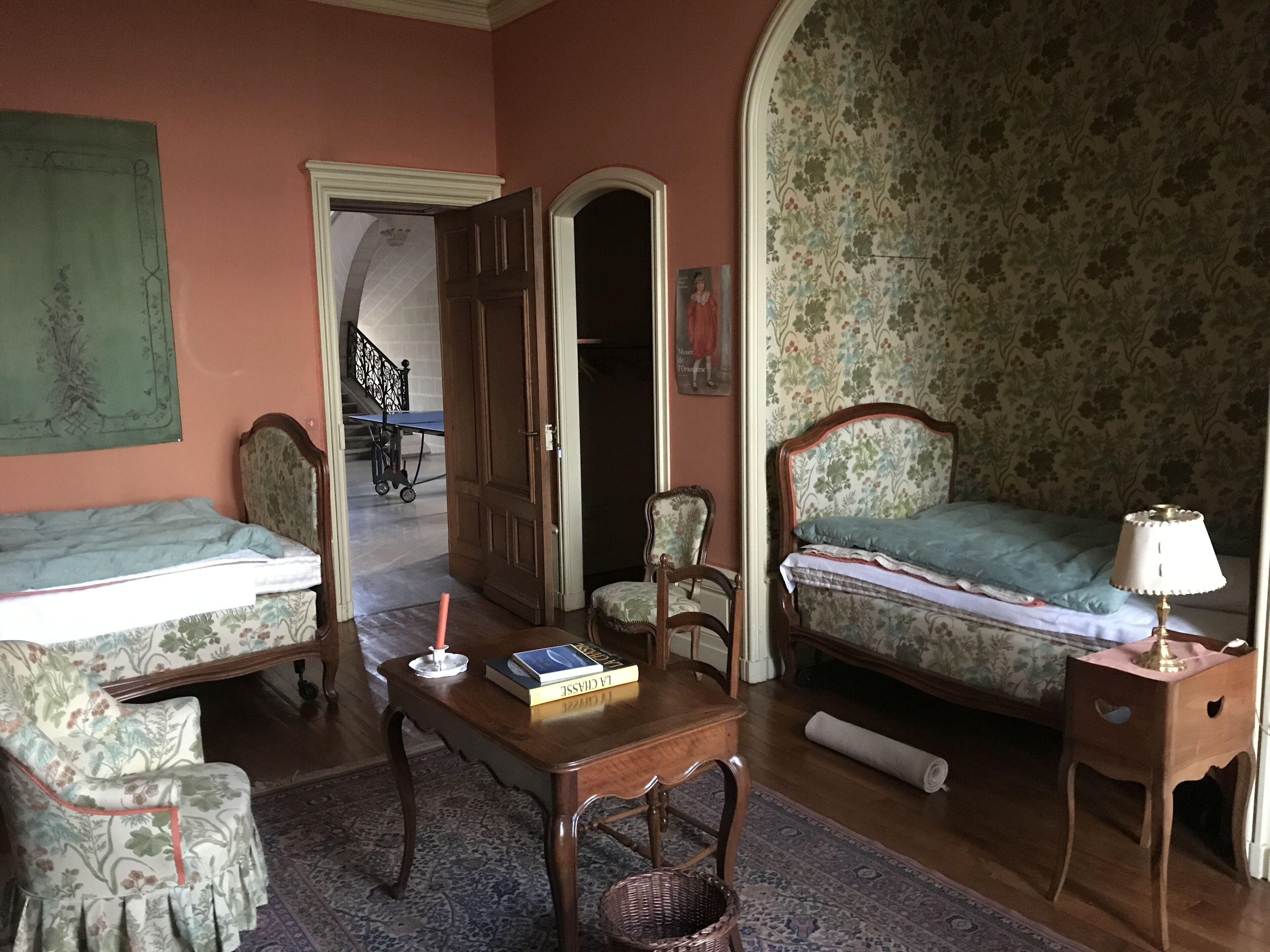 Une pièce comprenant un lit, une chaise, une table et une lampe. Une porte donne accès à une autre pièce.