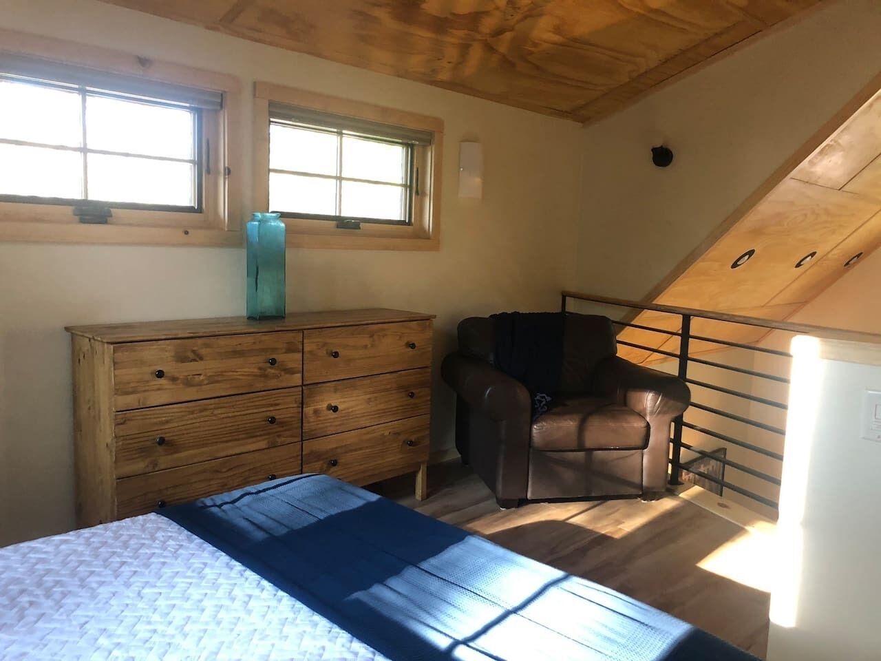 Ein Schlafzimmer mit Bett, Holzkommode und Stuhl. Zwei Fenster sorgen für natürliches Licht.