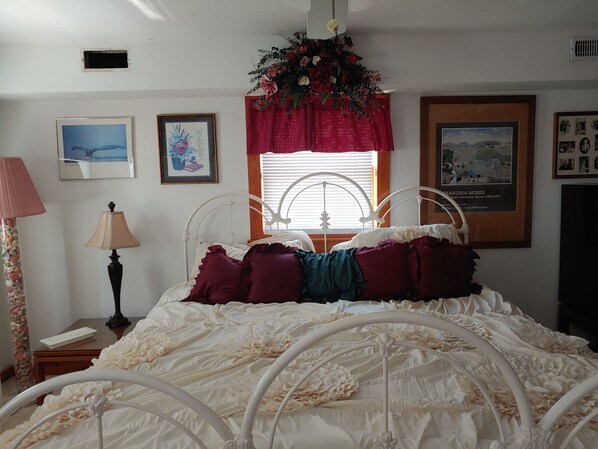 A bedroom with a white metal bed frame, a window with blinds, and a floral arrangement above the window. There are framed pictures on the wall and a lamp on a nightstand. - Beautiful Ocean Front Studio with direct beach and access to a deck. (Kill Devil Hills)