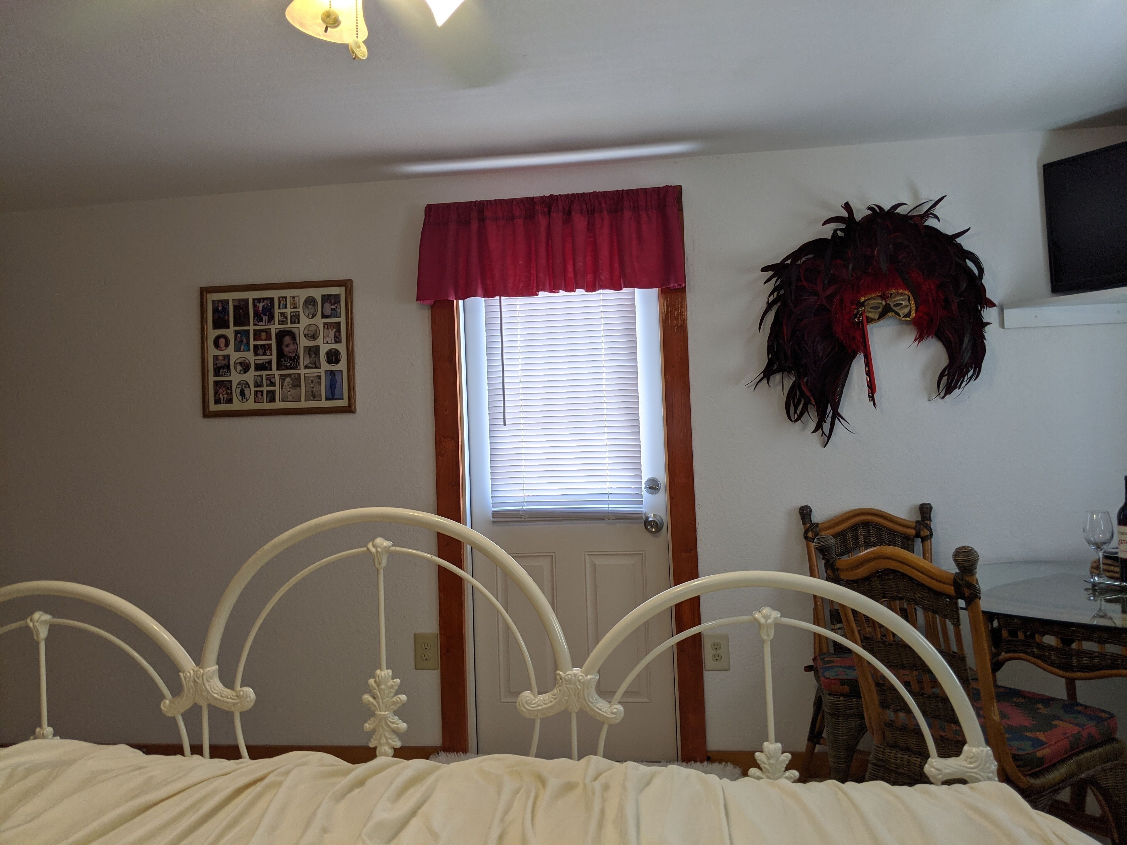 Ein Schlafzimmer mit weißem Metallbettgestell, einer Holztür, einem Fenster mit Jalousien, einer gerahmten Fotocollage und einer Wandmaske.