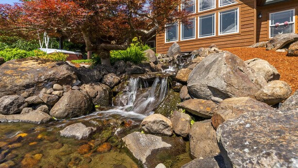 Een houten huis met een waterval, omgeven door rotsen en herfstkleurige bomen.