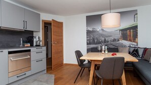 Une cuisine moderne dotée d’appareils électroménagers en acier inoxydable, un coin repas avec une table et des chaises en bois, et une grande fresque murale représentant un paysage montagneux.