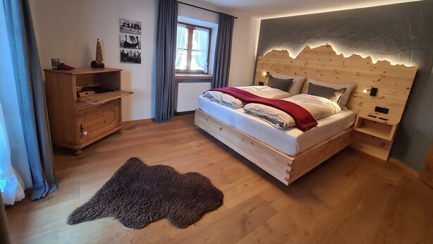 Une chambre avec un lit en bois, une table de chevet, une commode, une fenêtre avec des rideaux et un tapis sur le sol.