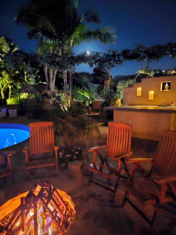 Un área junto a la piscina con sillas de madera, un hogar para la leña y una piscina pequeña.