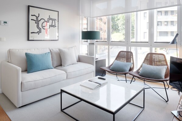 En moderne stue med en hvid sofa, to fletstole, et glasbord og et stort vindue med udsigt til bygninger.