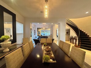 Uma sala de jantar moderna com uma mesa comprida, cadeiras estofadas, um detalhe decorativo central e uma escada que leva a um andar superior.