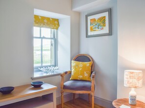 A room with a window, a chair with a floral cushion, a table with a bowl, a framed picture on the wall, and a lamp on a side table.