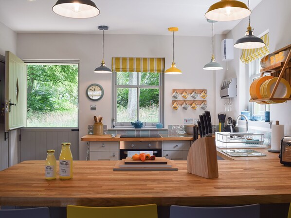 A modern kitchen with wooden countertops, hanging pendant lights, a window with a view of greenery, and a clock on the wall.