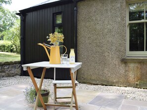 A table with a yellow watering can, a glass, and a bottle of apple juice outside a building.