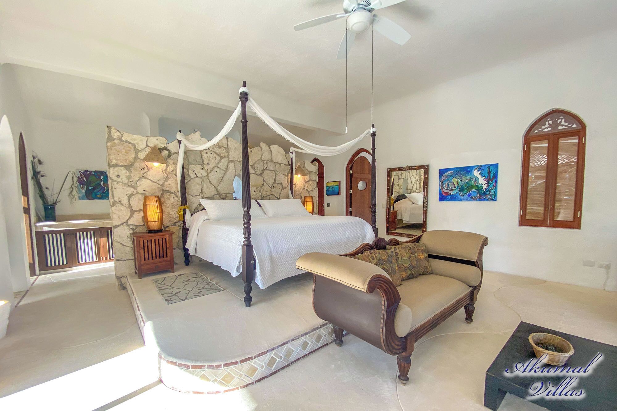A bedroom with a four-poster bed, a sitting area with a chair and a small table, stone walls, and artwork on the walls.