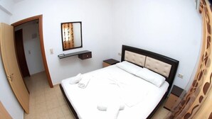 Una cama bien tendida con ropa de cama y toallas blancas, una mesita de noche, un espejo y una pequeña estantería.