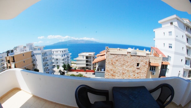 Un balcón con dos sillas y una mesa, con vistas a una ciudad costera con edificios y un cielo azul despejado.