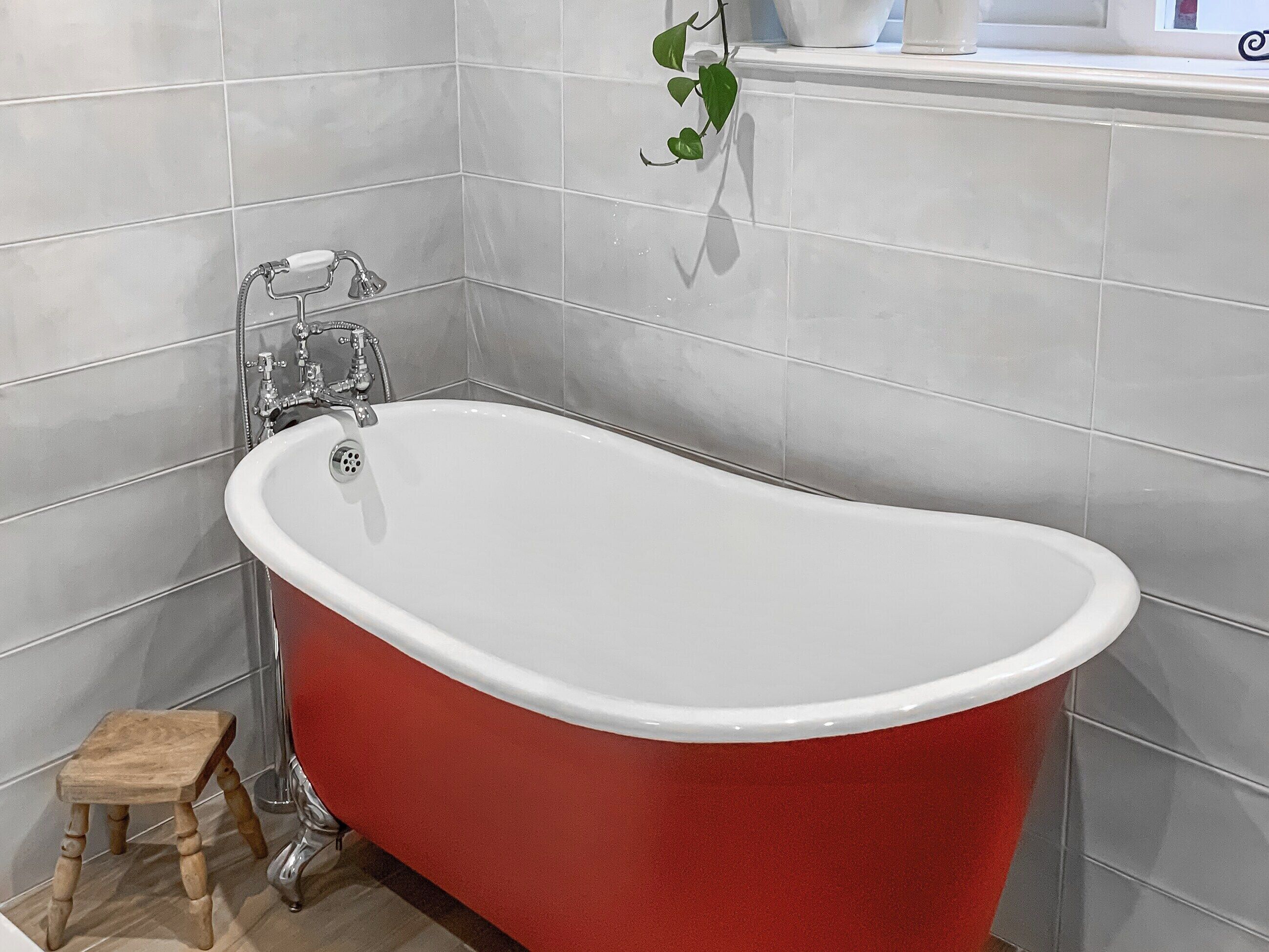 Une baignoire rouge, autoportante, dans une salle de bain aux murs recouverts de carreaux blancs et agrémentée d’un petit tabouret en bois.